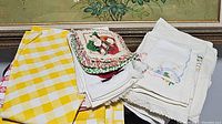 Folded linen textiles including yellow and white checkered cloth, holiday-themed pot holders, white linens with lace trim and embroidery.