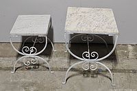 Front-facing view of pair of square garden tables with removable marble tops and white metal scrollwork bases on concrete floor.