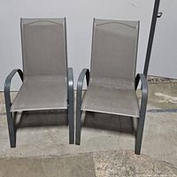 Pair of grey outdoor lounge chairs with mesh seating on dark metal frames, placed on concrete floor.