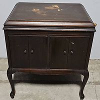 Closed view of dark mahogany phonograph console showing wear on top and classic design with scroll legs and pad feet.