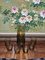 Full set of brown textured glass drinkware showing one pitcher and six glasses arranged in front of floral artwork.