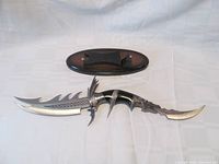 Full view of double bladed stainless steel decorative knife alongside its wooden display stand on white fabric background