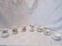 Six Royal Albert creamers and sugar bowls arranged side by side on fabric background. Varied floral patterns including violet, pink, and anemone petals visible.