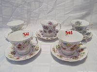 Five Royal Albert teacup and saucer sets arranged in a row on a light background. Two cups show Canadian flag emblem inside. Various floral patterns and gold rim accents are visible.