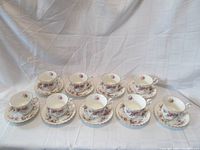 Full view of all 9 Royal Albert Lavender Rose teacup and saucer sets arranged in two rows on a draped light cloth.