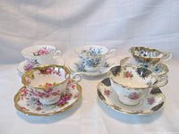 Five Royal Albert teacups and saucers arranged on white fabric background showing variety of patterns including floral and gold lattice accents.