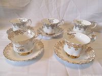 Five porcelain teacups and saucers with decorative gold floral patterns and gold trim, arranged on white fabric background.