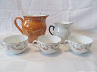 Front view of three bridal rose porcelain cups and two vintage pitchers, one in rainbow lustre and one brushed orange.