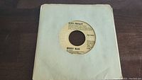 Two images of a 45rpm vinyl record by Elvis Presley with Moody Blue on Victor label in plain white paper sleeve, slightly worn.