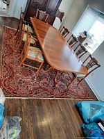 Antique Persian area rug placed under a wooden dining table with multiple chairs around it. Rug shows traditional Persian patterns with red dominant colors, and evident wear.