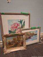 Three framed artworks arranged on the floor: pink flower watercolour in light wood frame, landscape watercolour in white frame, autumn landscape oil painting in ornate frame