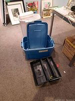 Four plastic bins and toolbox displayed on carpet. Blue bin with lid, white bin in background, black toolbox with compartments in front.