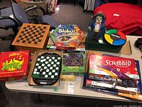 Top view of multiple boxed board games and game items including Apples to Apples, Blokus Trigon, Scrabble, pirate figure, Simon game, and a wooden chess/checkerboard.