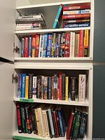 Four shelves filled with a large collection of fiction books with titles and author names clearly visible. Shelves contain multiple paperbacks and some hardcover books.