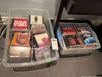 Two plastic bins filled with assorted pop and rock music CDs including notable titles like The Rolling Stones and Linkin Park.