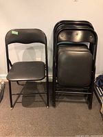 Five black folding chairs stacked against a wall showing metal frames and vinyl padded seats and backs.