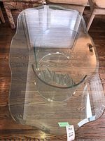 Top-down angle showing entire glass coffee table with curved, bevelled top edges, circular lower glass shelf, and curved glass legs on wood floor background.