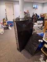 Photo showing metal wire dog crate folded with black plastic tray and quilted black bed insert leaning against it, in basement setting.