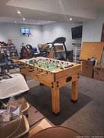 Full view of the foosball table in a basement room with visible wood base and legs, basketball hoop, treadmill, boxes and other items in background.