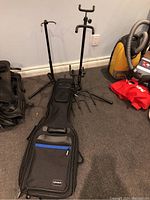 Photo of three black metal guitar stands positioned on carpeted floor along with a black soft guitar case with blue accent pocket.