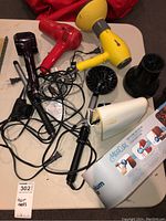 Photo showing multiple hair dryers in yellow, red, and black colors, curling tools including wand and attachments placed on a table with tangled cords.
