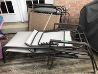 Four beige fabric lounge chairs with black metal frames stacked and set on a wooden deck, showing two less weathered and two more weathered chairs.