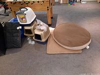 Four covered litter boxes in white and beige plastic with front entry flaps, two round brown dog beds with rectangular mats beside a white plastic water dispenser. Items arranged on a carpeted floor with a foosball table in background.
