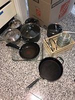 Photo showing assorted black cooking pots with glass and metal lids, non-stick frying pans, and glass bowls stacked in a wooden rack.