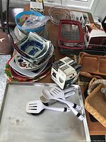 View of assorted bowls stacked, metal wire basket, 3-tier buffet set box, Auberge fondue chocolate set box, wooden serving trays, kitchen utensils, and a rectangular metal tray.