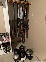 Wide view of skis on a wall rack and helmets on the floor below, also ski poles and some ski boots visible.