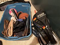 Bin containing assorted baseball gloves, bat, basketball, knee pads, black protective gear and some balls, next to tennis racket covers labeled Prince and Dunlop, and a Head racket cover