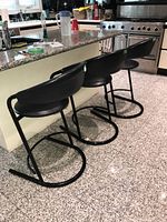 Photo showing three of the four black kitchen stools lined up next to a kitchen island, showing curved backrests, round seats and black metal circular bases.