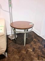 Side view of round wood top side table showing metal legs and base ring, with lamp and couch nearby.
