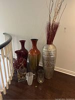 Wide view of all six decorative vases grouped on dark wood floor near staircase showing size and variety.
