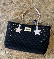 Front view of a black quilted Betsey Johnson handbag decorated with gold star studs and pink star-shaped charms on chain handles laid flat on a wooden surface.