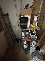 Metal wire display racks filled with multiple paint cans, fabric rolls, and painting supplies stored in basement corner.