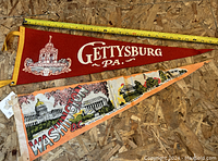 Two vintage pennants laid out on wood surface with a measuring tape showing size, one red for Gettysburg PA, one cream for Washington D.C.