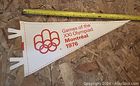 White leatherette triangular pennant with red print showing the Olympic rings and text 'Games of the XXI Olympiad Montreal 1976' placed on a plywood surface with measuring tape above it.