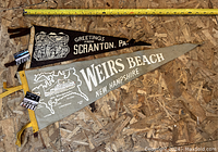 Photo of two vintage souvenir pennants lying flat on a wood surface, one black for Scranton PA and one gray for Weirs Beach NH with map detail.