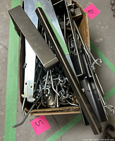 Box with assorted metal brackets, hooks, and hangers in silver and black finishes piled together showing variety in sizes and shapes.