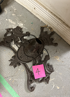 Dark cast iron vintage flag holder with coat of arms design, shown laying on floor with pink sticker marked '4'. Central angled circular socket for flagpole visible.
