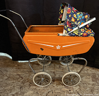 Full side view of the vintage orange metal child's toy pram with floral fabric canopy attached, showing the metal frame and wheels.