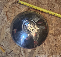Top view of a round metallic Volkswagen hubcap placed on plywood with a measuring tape showing about 6 inches diameter. The hubcap has embossed VW logo in center, reflective surface with some minor wear consistent with vintage condition.