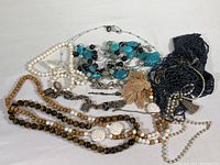 Overview of assorted necklaces and bracelets showing different color beads and materials including white, turquoise, black, brown, and metal chain links with tassels.