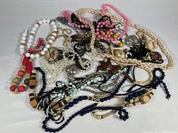 Various costume jewelry necklaces in white, pink, wood, faux pearls, and metal pendants piled together on a white surface.
