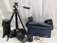 Full kit showing camera body with zoom lens mounted, additional 50mm lens, shutter release cable, small tripod, and Mustang camera bag.