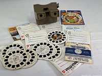 Beige View-Master viewer with seven reels and accompanying sleeves including Disney on Parade and Happy Days. Various titles visible on reel inserts. All items displayed on white surface.
