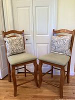 Two dark honey colored wood counter stools with plaid upholstered seats and decorative cushions in front of white doors on wood floor.