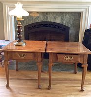 Two wooden side tables with brass drawer pulls and carved leg details placed side by side, with vintage-style wood and brass lamp on one table.