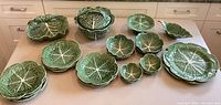 Full view of all cabbage leaf-designed ceramic dishes arranged on table, shows variety of plates, bowls and serving pieces.